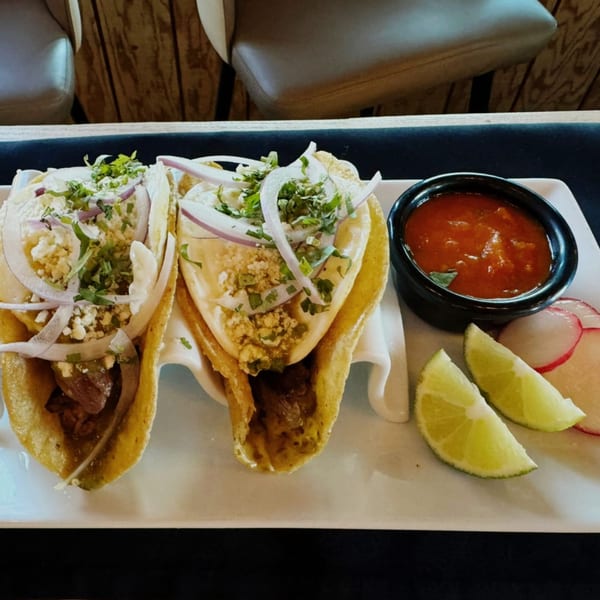 Two IXCHEL Mexican Cuisine tacos with onions, cilantro, and cheese, served with salsa, lime, and radish.