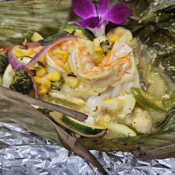 Shrimp, corn, and vegetables in a creamy sauce, garnished with a purple orchid, served in a banana leaf.