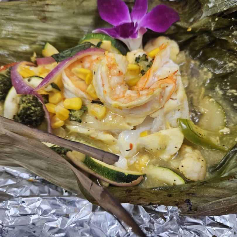 Shrimp, corn, and vegetables in a creamy sauce, garnished with a purple orchid, served in a banana leaf.