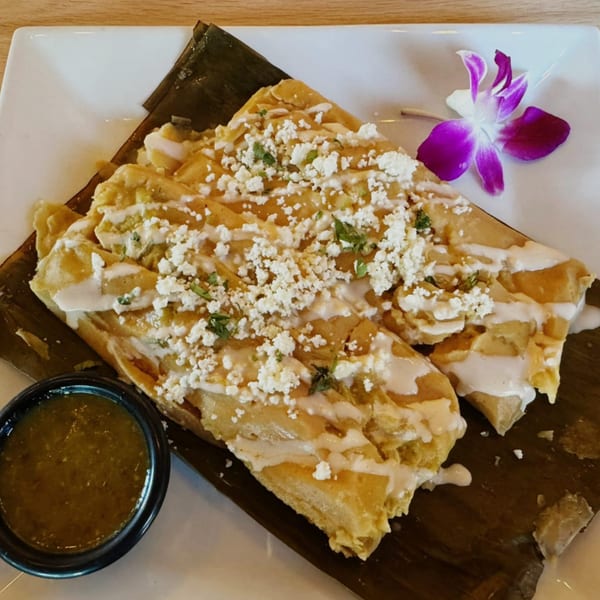 Two tamales topped with white sauce, crumbled cheese, and herbs, served with green salsa and an orchid.