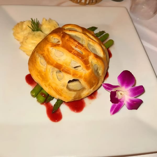 Golden pastry dish with asparagus, mashed potatoes, red sauce, and a purple orchid on a white plate.