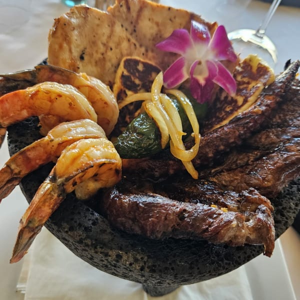 A molcajete filled with grilled shrimp, steak, cheese, onions, peppers, and a purple orchid.