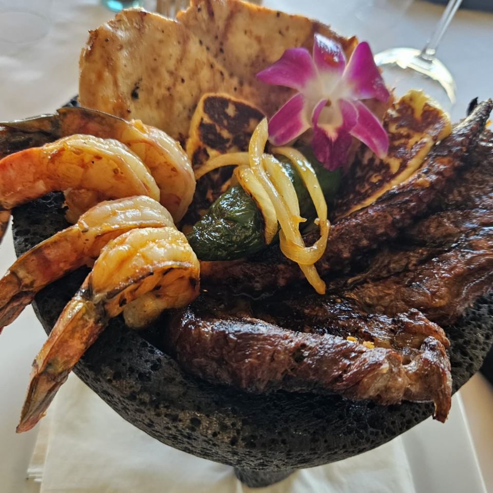 A molcajete filled with grilled shrimp, steak, cheese, onions, peppers, and a purple orchid.
