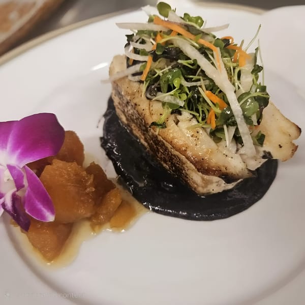 Grilled fish with black sauce, topped with microgreens and shredded vegetables, next to a purple orchid and fruit.