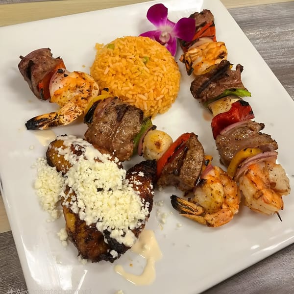 Grilled shrimp and beef skewers with rice, plantains, and a purple orchid on a white plate.