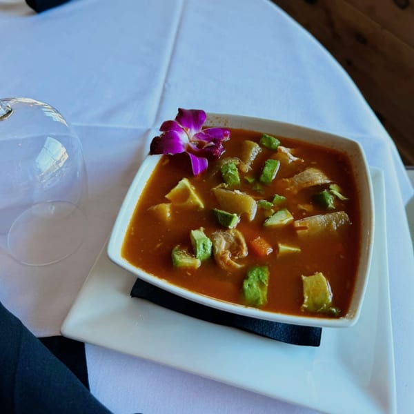 A bowl of Mexican soup with avocado and meat, garnished with a purple orchid, on a white tablecloth.