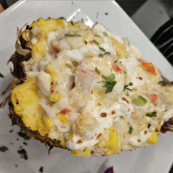Half a pineapple filled with melted cheese, corn, peppers, and cilantro on a white plate.